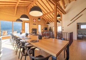 A spacious dining area with a long wooden table and black chairs, wooden beams on the ceiling, and large windows offering a view of the outdoors.