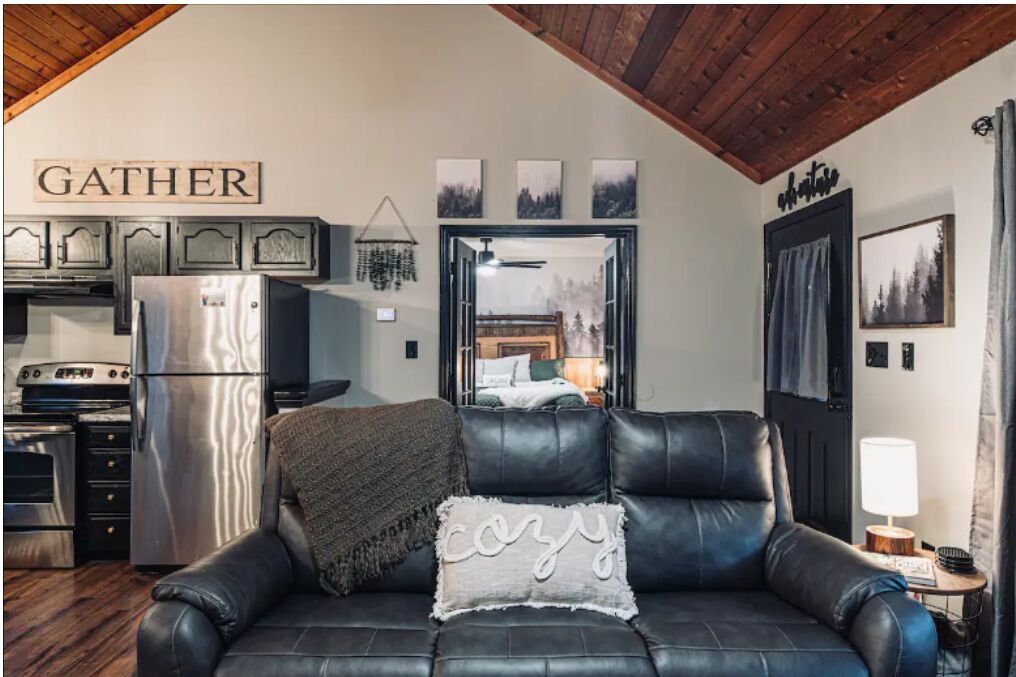 A cozy living room with a black leather sofa, a 'GATHER' sign, a refrigerator, and a wooden ceiling.