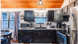 Una cocina con techo de madera, gabinetes negros, electrodomésticos de acero inoxidable y una ventana con cortinas.