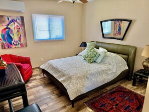 A bedroom with a bed, a red chair, a wooden floor, a ceiling fan, and a window with blinds. - Suite Stay with Heavenly Views (Tucson)