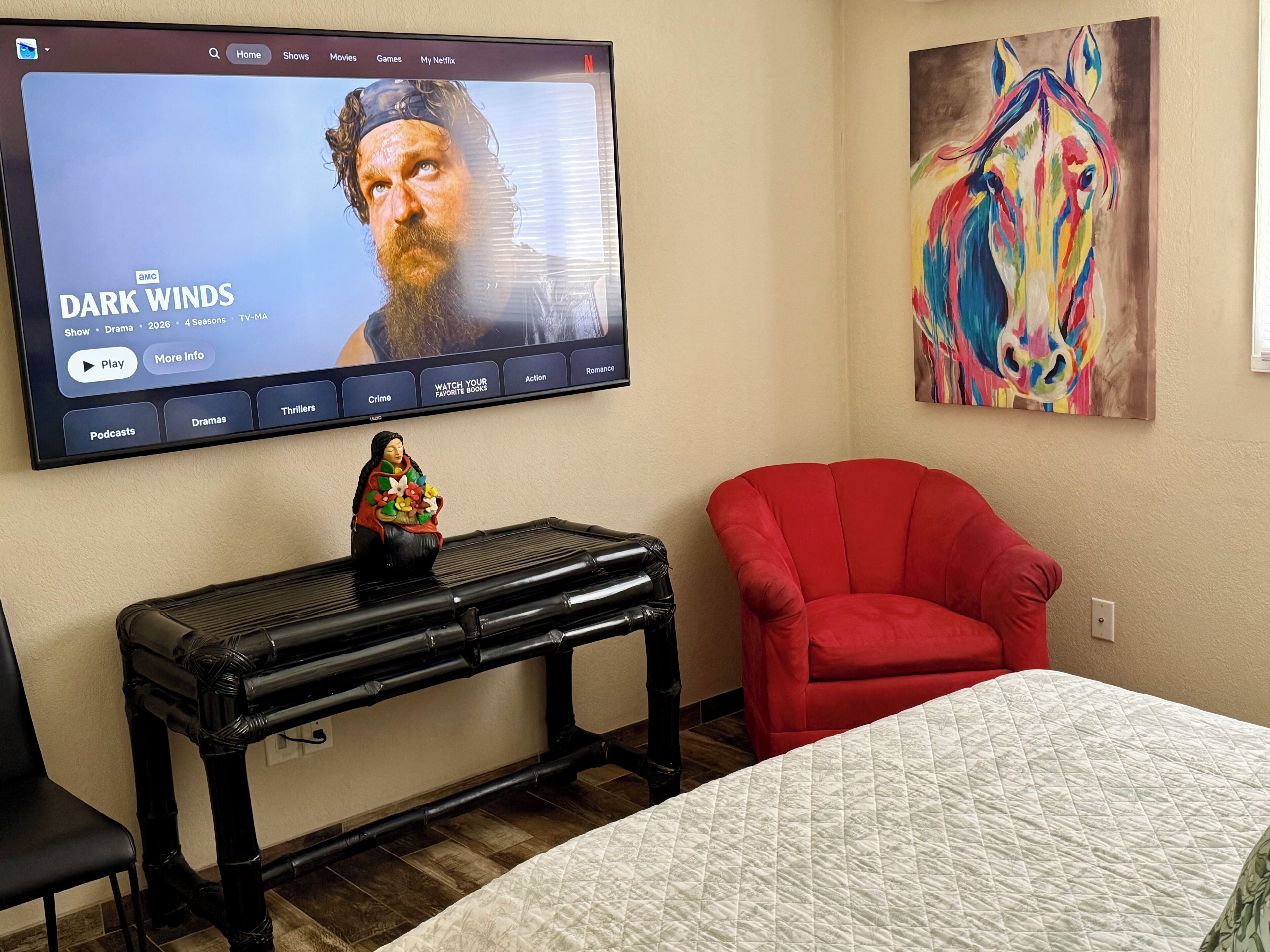 Ein Zimmer mit einem Bett, einem roten Sessel, einem Fernseher, der 'DARK WINDS' zeigt, und einem farbenfrohen Gemälde an der Wand.