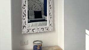 A bedroom with a white ornate mirror, a bed with a black headboard, and a small wooden table with a blue can. - Pharus miseni (Bacoli)