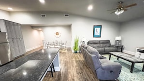 Eine moderne Küche mit dunkler Arbeitsplatte und Holzboden, ein graues Ecksofa, ein Couchtisch und ein Deckenventilator.