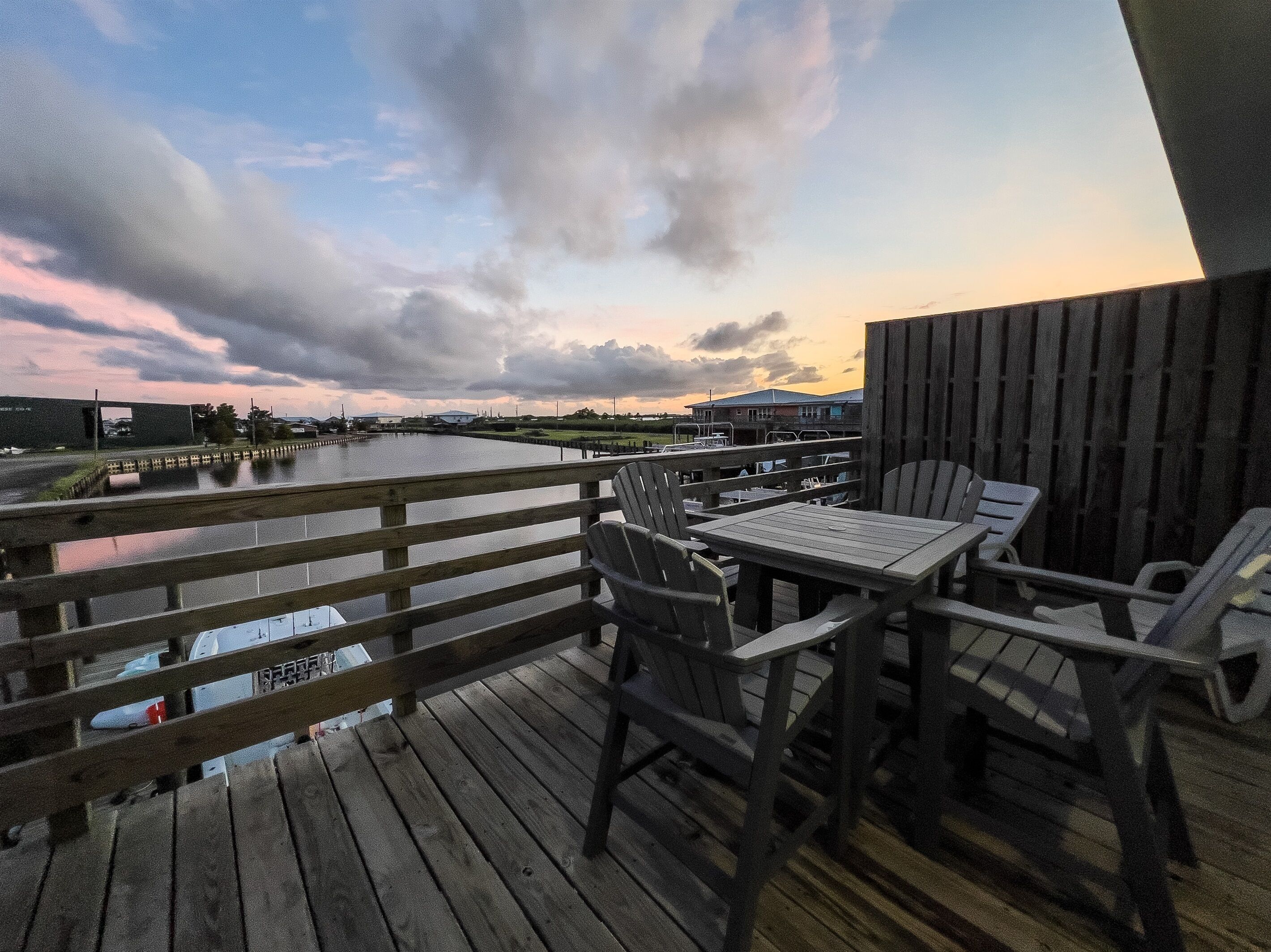 Une terrasse en bois, agrémentée de tables et de chaises, qui offre une vue sur une étendue d’eau, avec des bâtiments en arrière-plan.