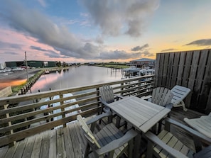 Een houten terras met tafels en stoelen met uitzicht op kalm water, met een steiger en gebouwen op de achtergrond.