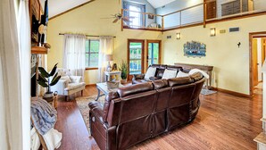 A spacious living room with a brown leather sofa, a white armchair, and a coffee table. A staircase with wooden railings leads to an upper level.