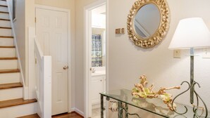 A hallway with a white door, a staircase, a wall-mounted mirror, and a glass table with decorative items.