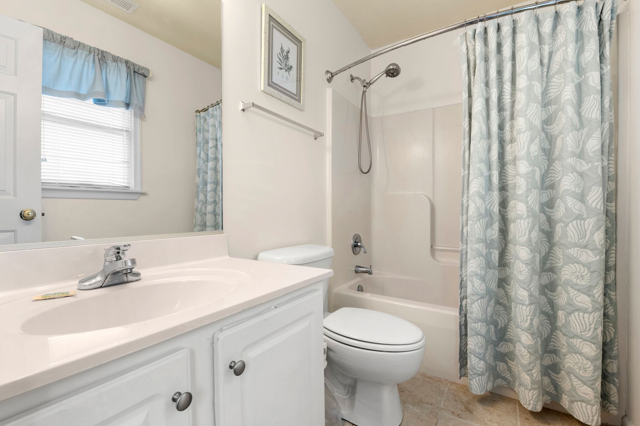 Une salle de bain avec un lavabo, des toilettes et une baignoire, tous de couleur blanche. On y trouve un rideau de douche orné de motifs de feuilles, une fenêtre avec des stores et un tableau encadré accroché au mur.