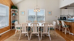 A dining area with a wooden ceiling, white walls, and a large window. There is a table with white chairs and a hanging light fixture.