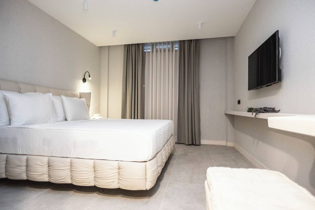 Moderni hotellihuone, jossa on suuri sänky, yöpöytä, seinälle asennettu taulutelevisio ja ikkuna, jossa on verhot.