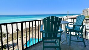 A balcony with two green Adirondack chairs and a table, overlooking a beach with a clear blue sky.