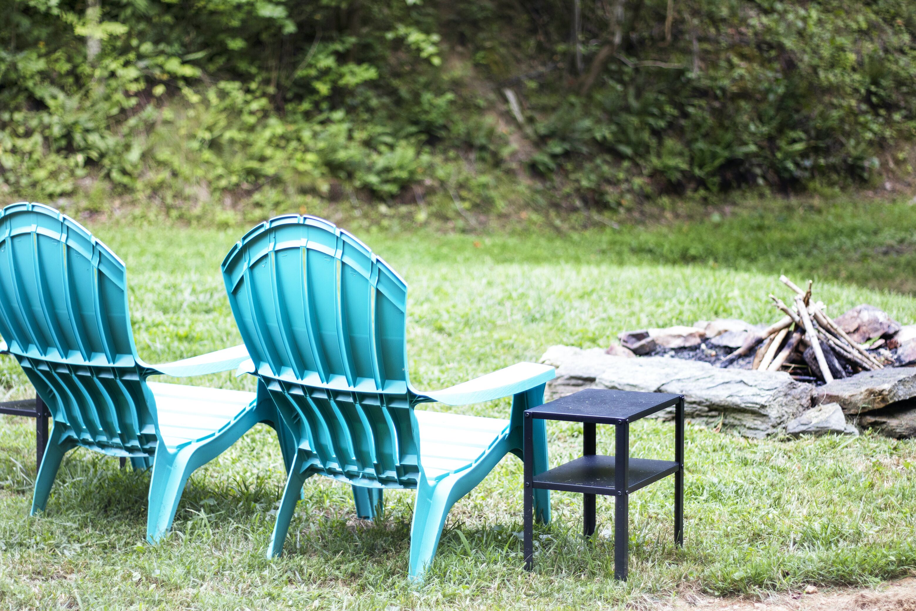 Two turquoise Adirondack chairs and a small black table in a grassy outdoor area with a pile of logs and rocks in the background.