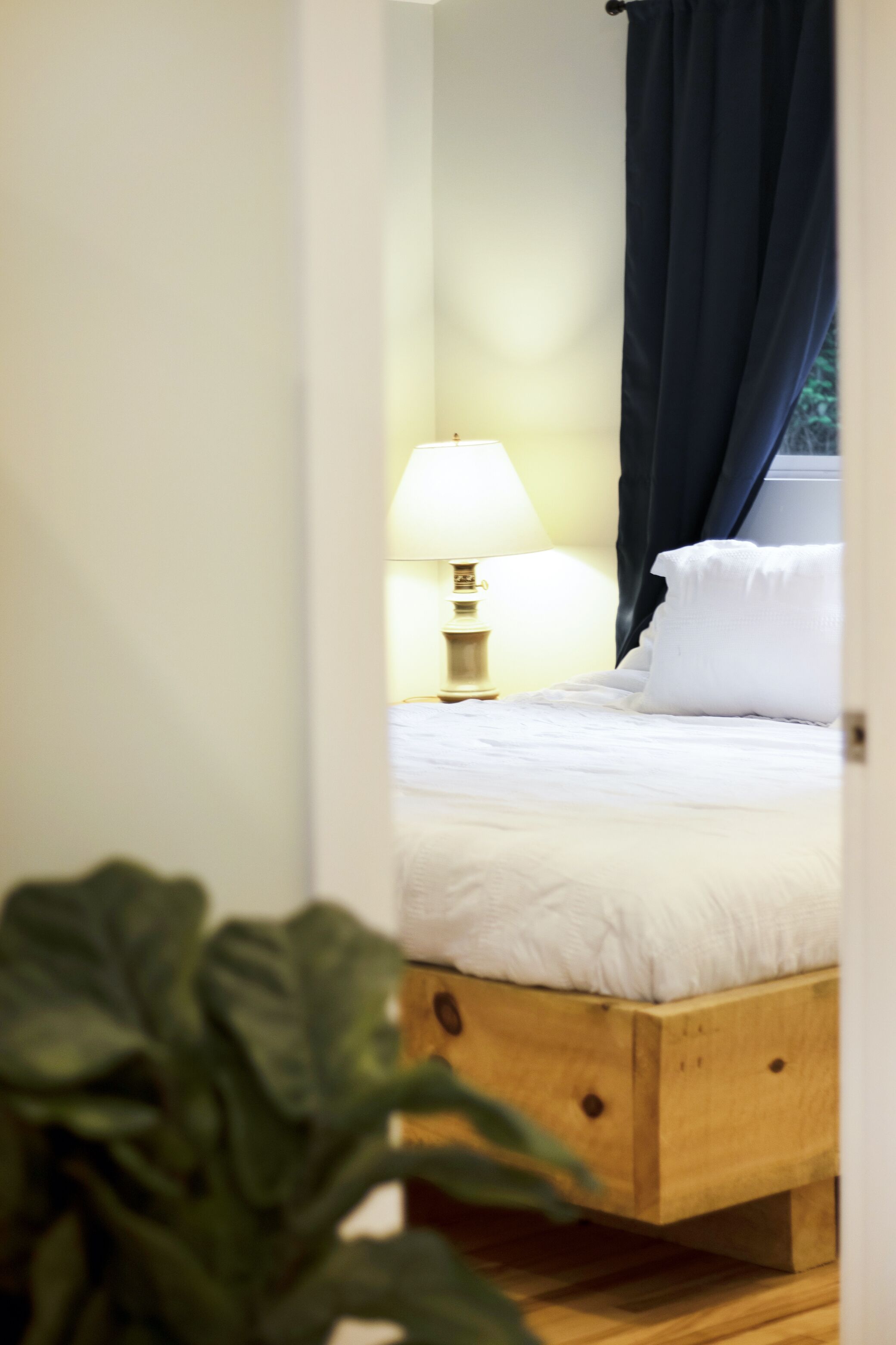 Un dormitorio con una cama de madera, ropa de cama blanca, una lámpara de noche y una ventana con cortinas oscuras.