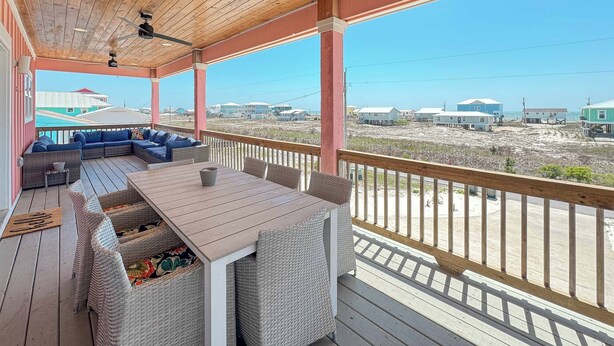 A covered outdoor area with a wooden deck, wicker furniture, and a view of a beach and buildings in the distance. - Your Private Paradise Awaits! (Gulf Shores)