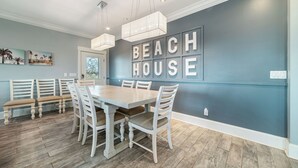 A dining area with a large wooden table, white chairs, and a bench. The wall behind features a large sign that reads "BEACH HOUSE." - Your Private Paradise Awaits! (Gulf Shores)