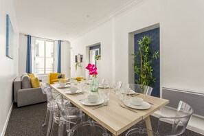 Un coin repas avec une table en bois dressée pour six personnes, des chaises en plastique transparent, un canapé avec des coussins jaunes et une grande fenêtre ornée de rideaux bleus.