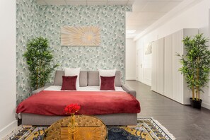 A modern bedroom with a bed, a red throw, and a vase with red flowers. There are two potted plants on either side of the bed.