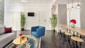 A modern living room with a sofa, a blue armchair, a coffee table with a vase of red flowers, a dining area with a wooden table and bar stools, and two potted plants. - Merveil - Family Suite - Marais - Sebastopol III (Paris)