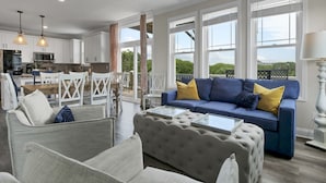 Amplia sala de estar con sofá azul, sillas blancas y una mesa de comedor. Se ve una cocina al fondo y grandes ventanales que dejan entrar luz natural.