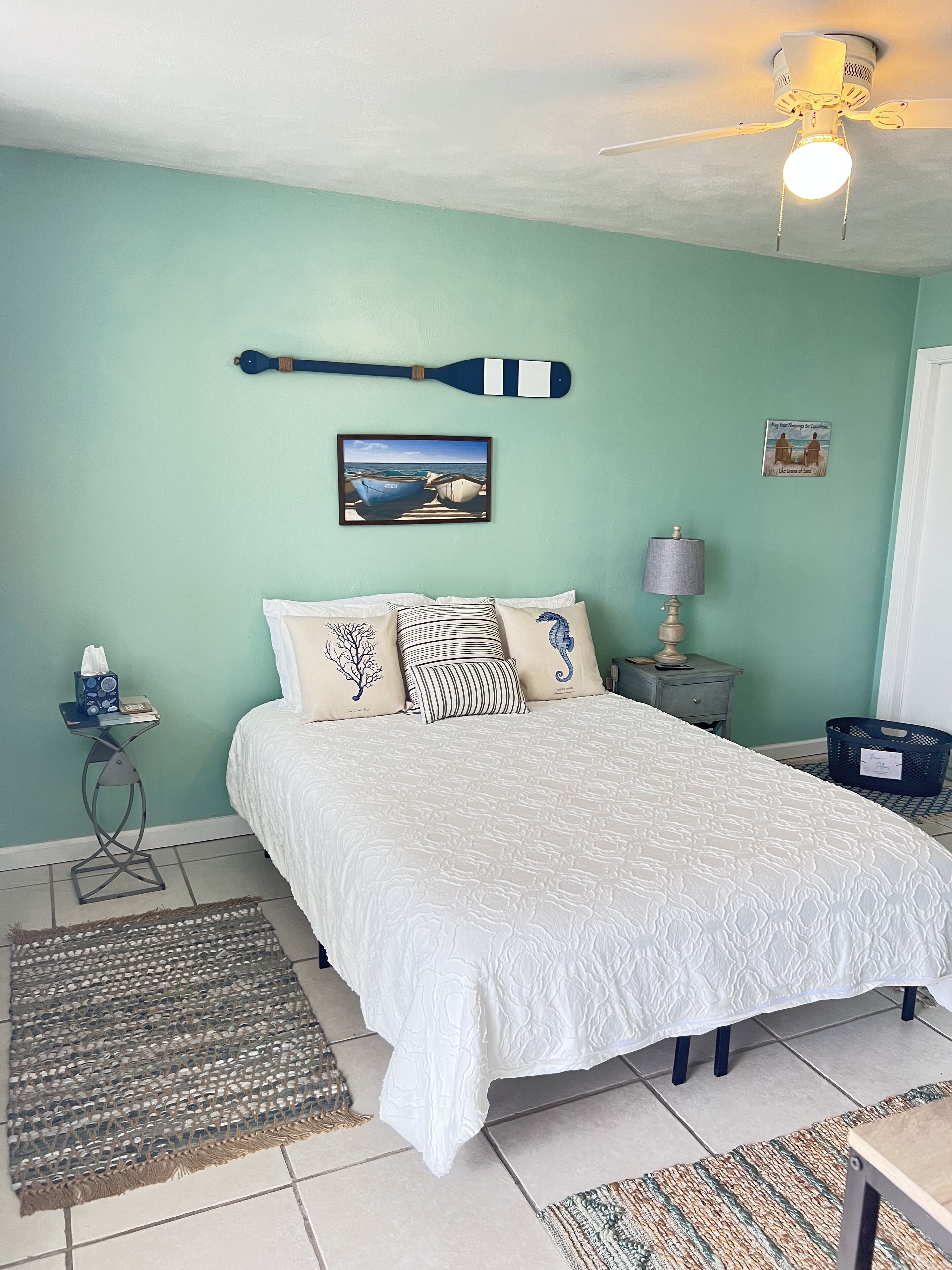 Ein Schlafzimmer mit Bett, Kissen, einem gerahmten Bild, einem kleinen Tisch, einer Lampe und einem Deckenventilator.
