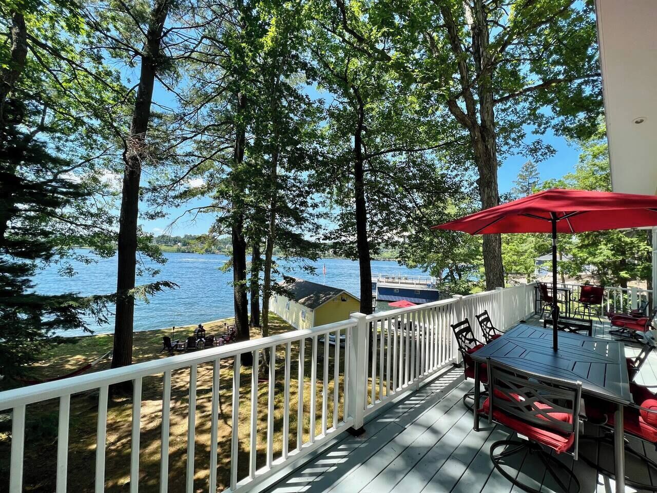 A deck with a table and chairs, overlooking a lake with trees and a small building.