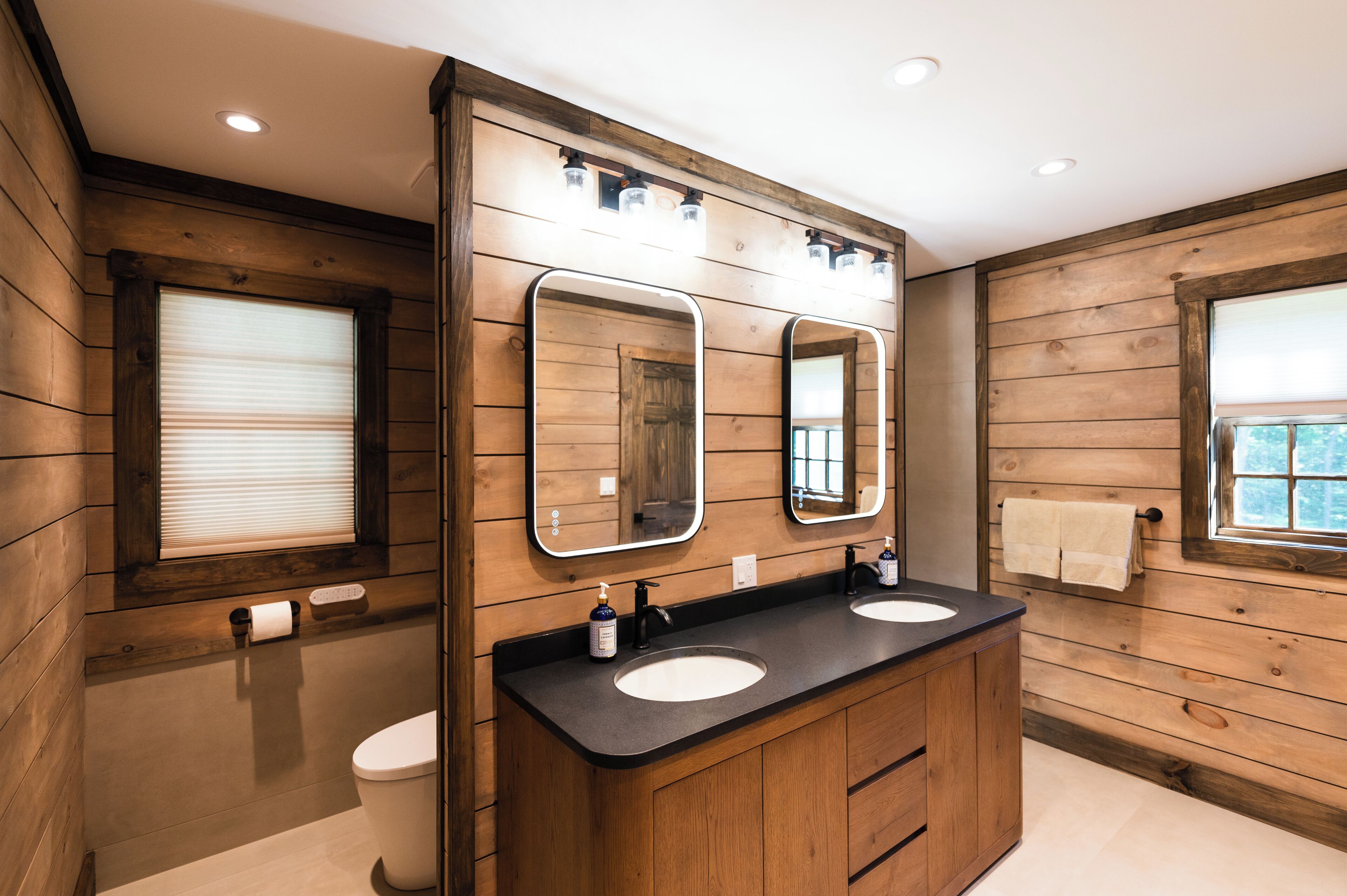 Une salle de bain de style rustique, avec des murs en bois, deux lavabos, des toilettes et un grand miroir.