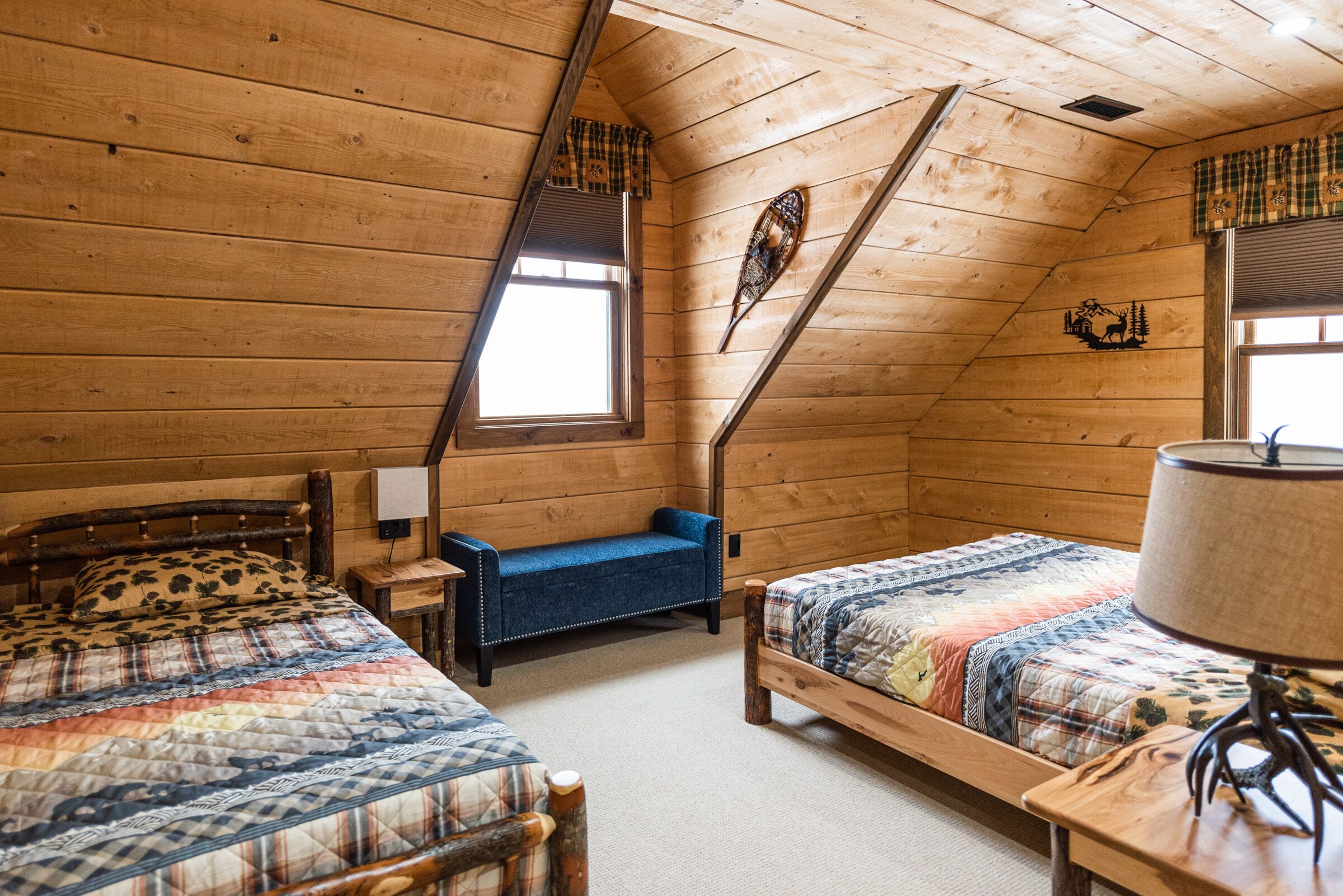 A cozy cabin room with two wooden beds, a bench, a lamp, and a window with plaid curtains.