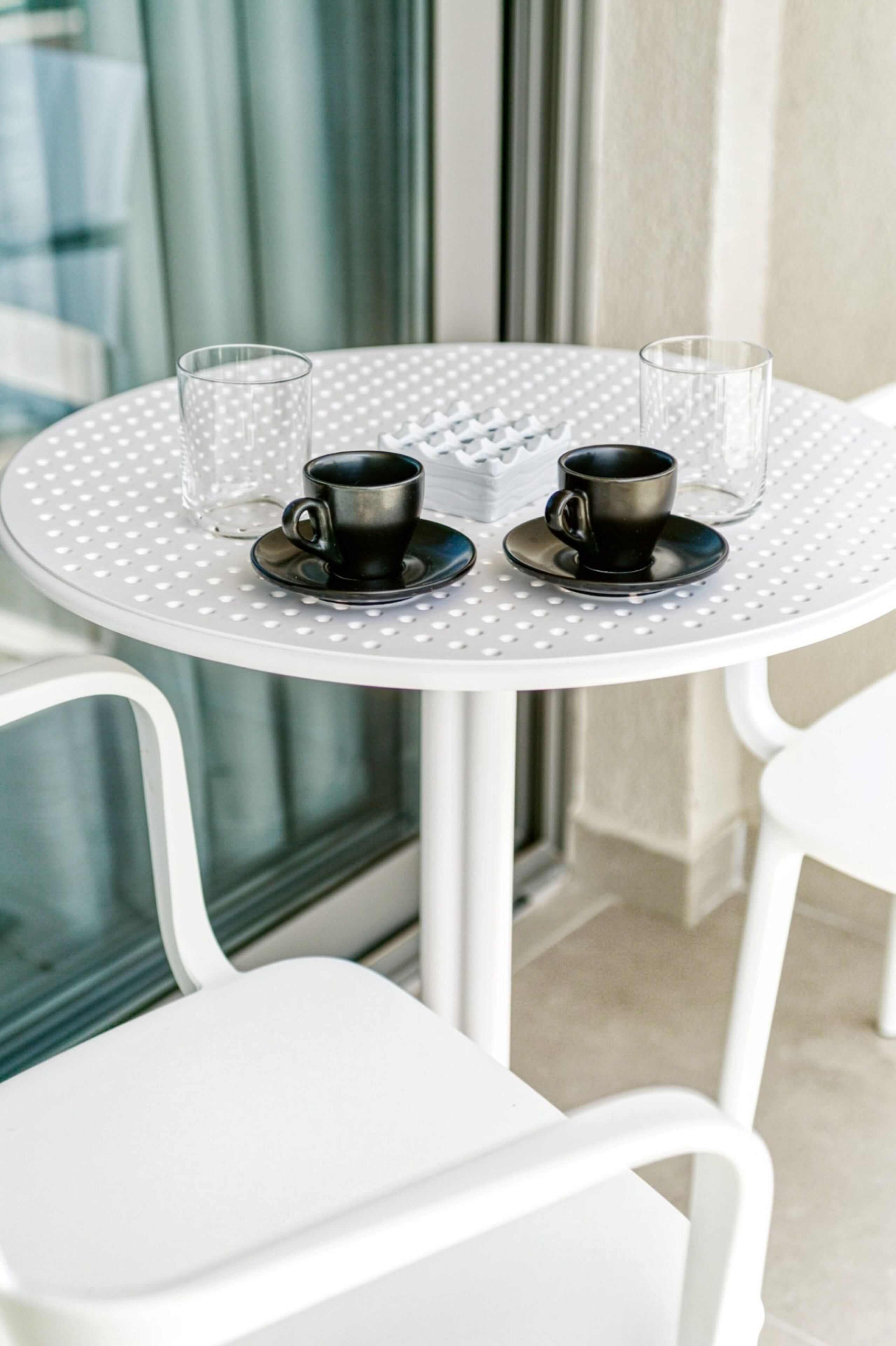 Una mesa redonda con dos tazas y platillos negros, y dos vasos de cristal, sobre una mesa blanca con diseño a lunares.
