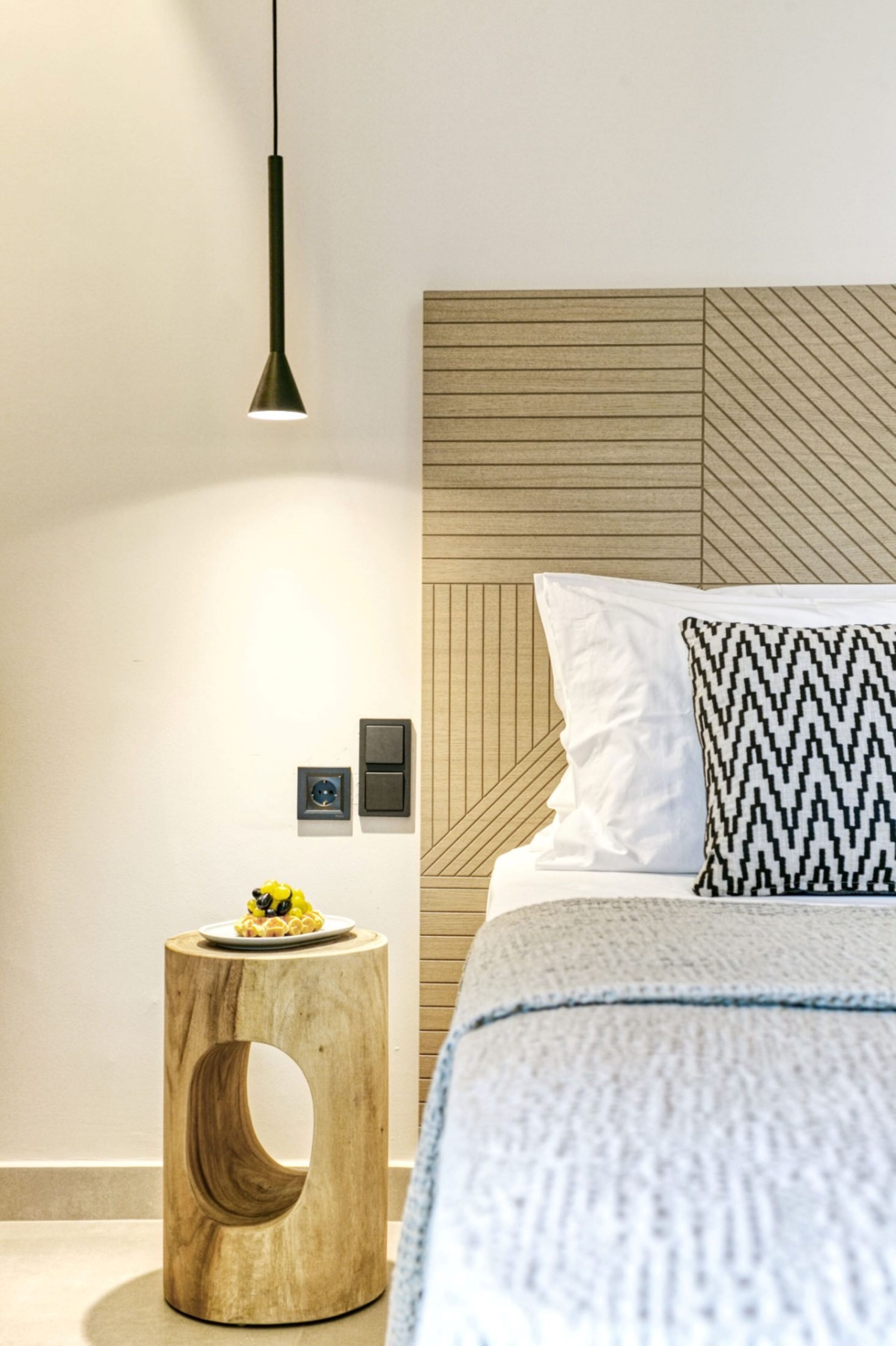 Un dormitorio moderno con cabecera de madera, un taburete redondo también de madera con una bandeja de fruta, una lámpara colgante y una cama con un cojín estampado.