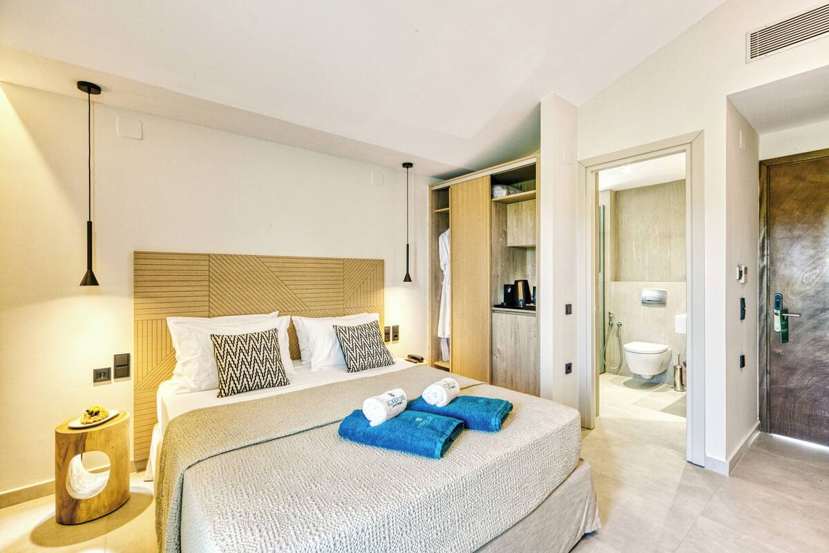 Quarto de hotel moderno com uma cama grande, cabeceira de madeira, mesa de cabeceira e um banheiro visível por uma porta aberta.