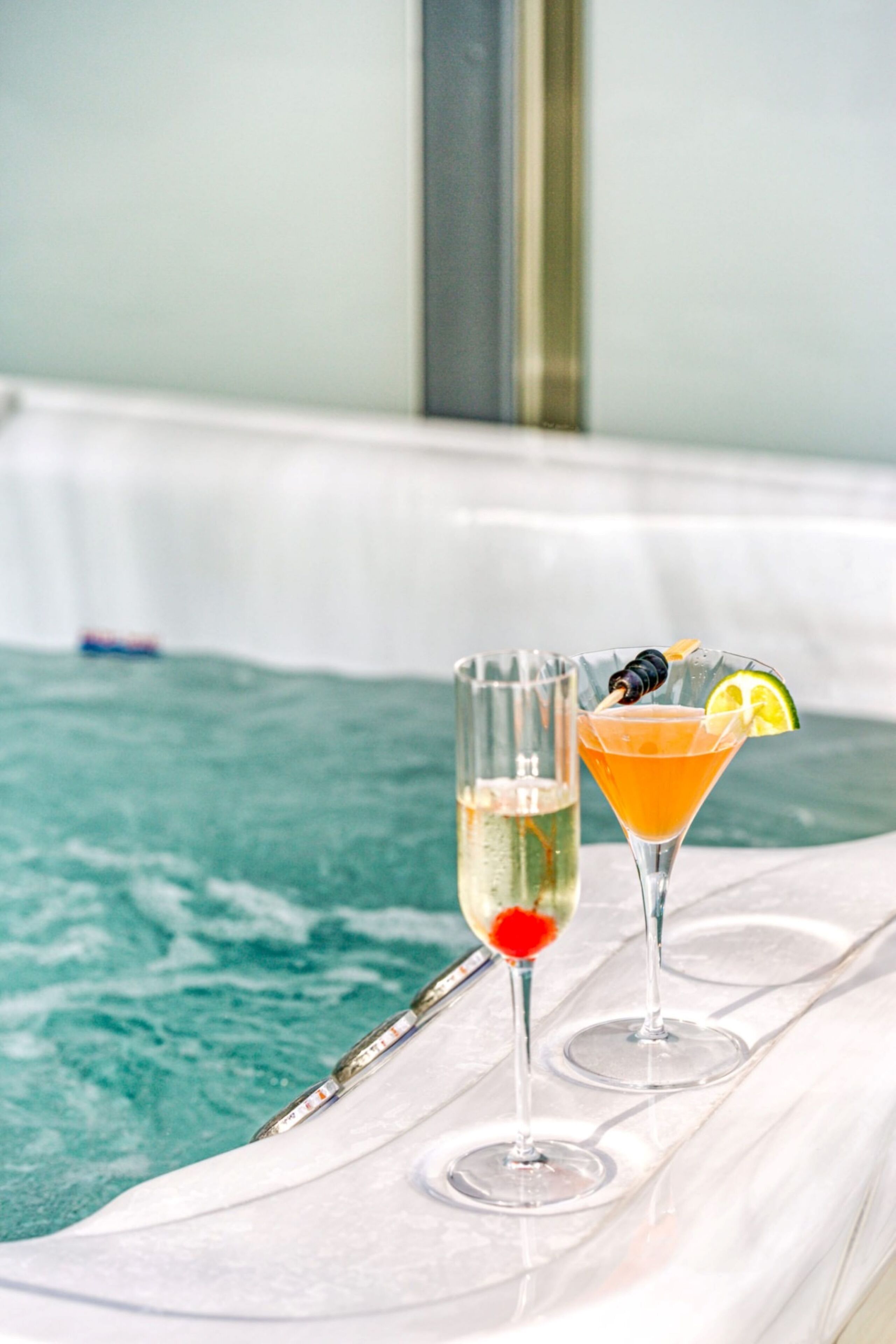 Two glasses of beverages on a white tray by a pool with a blue water surface.