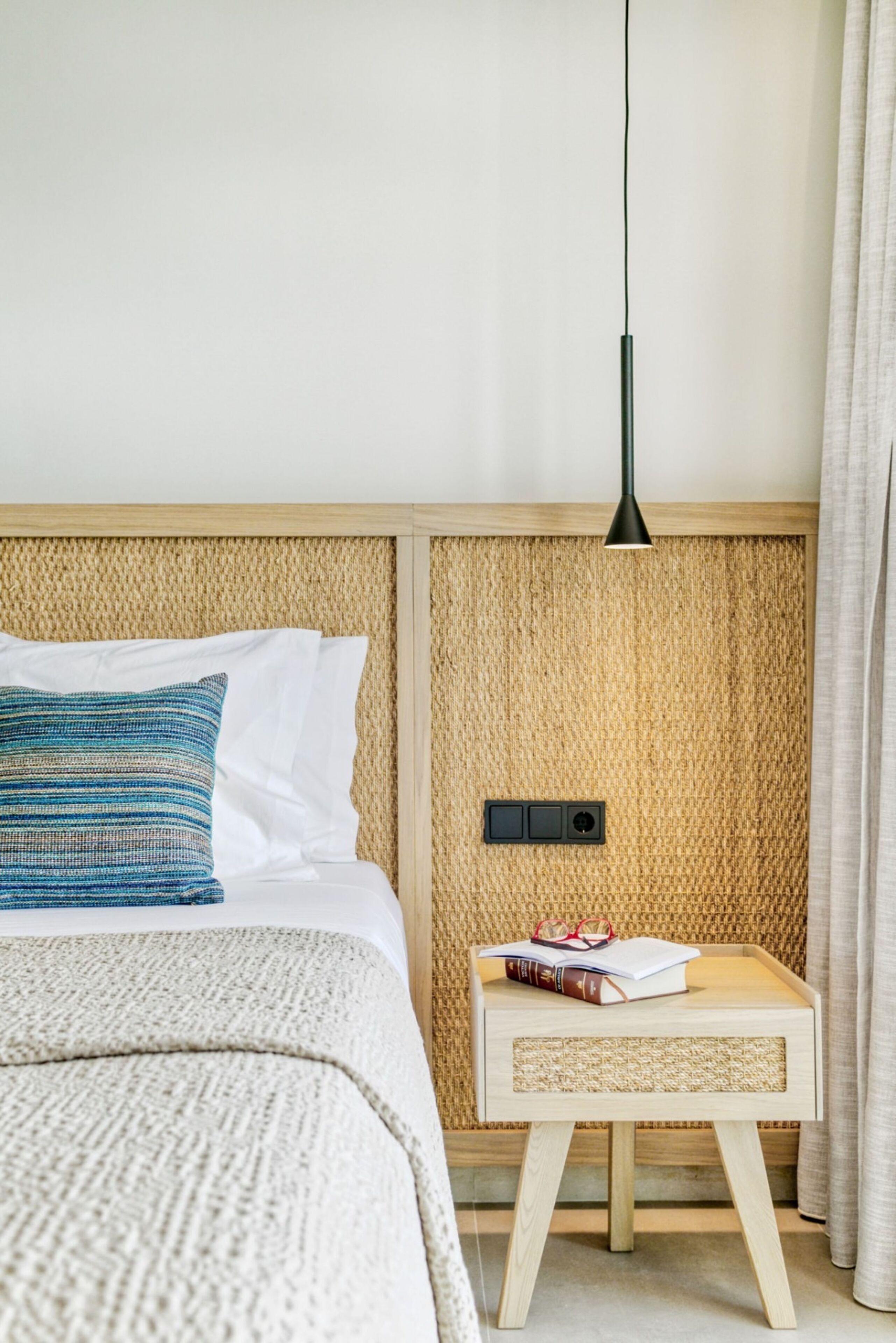 Een netjes opgemaakt bed met een gestreept kussen, een nachtkastje met een boek en een bril, en een wandlamp.