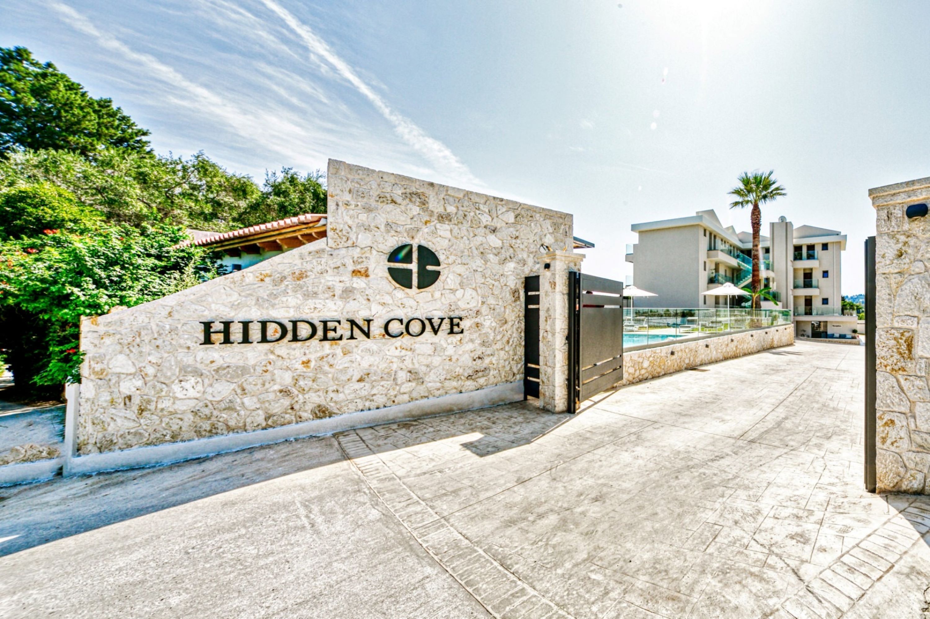 Ein Steineingang mit der Beschilderung "HIDDEN COVE", der zu einem modernen Gebäude mit Pool und einer Palme führt.