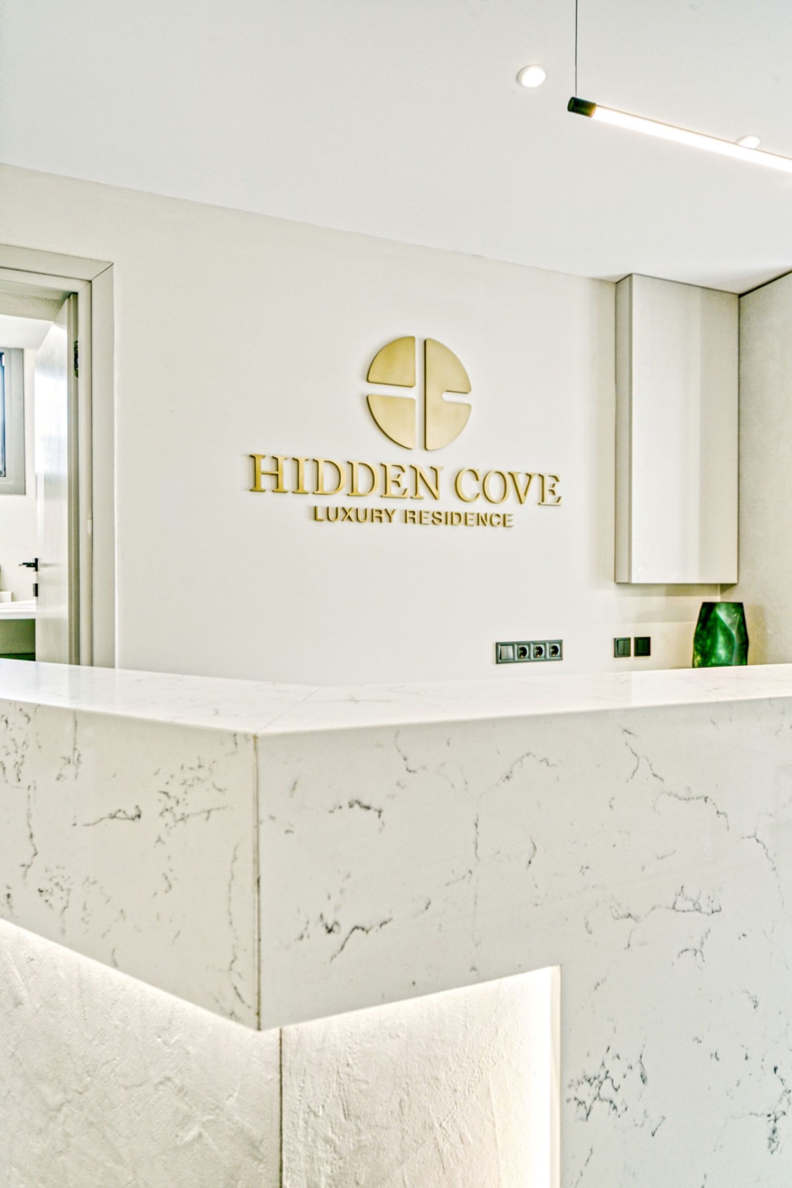 A reception area with a marble counter, a wall-mounted sign reading "Hidden Cove Luxury Residence," and a modern light fixture.