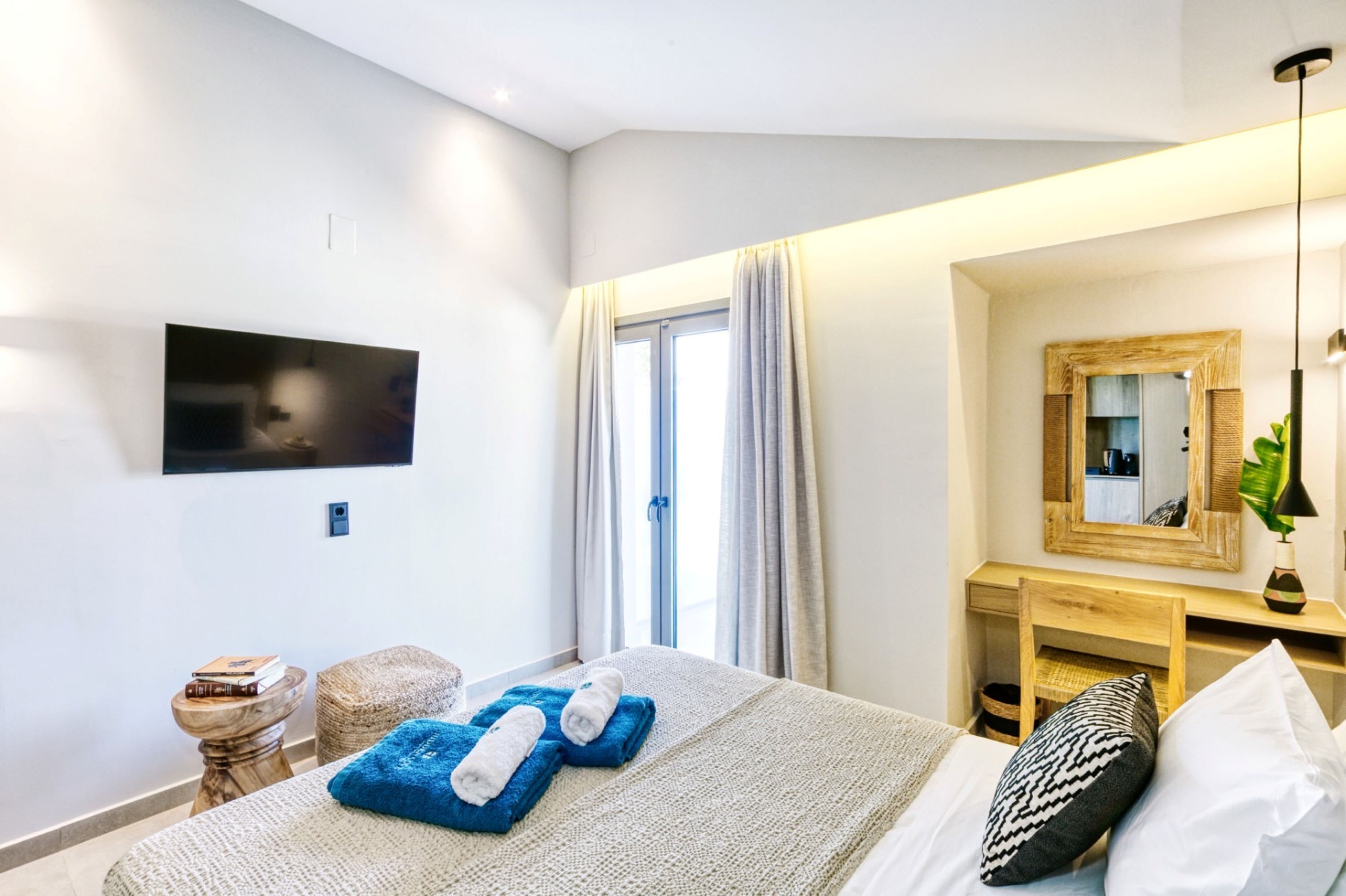 A modern hotel room with a bed, a TV mounted on the wall, a wooden desk, and a large window with curtains.