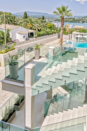 A modern outdoor terrace with glass railings, a pool, and a view of the surrounding landscape. - Hidden Cove Corfu (Corfu)