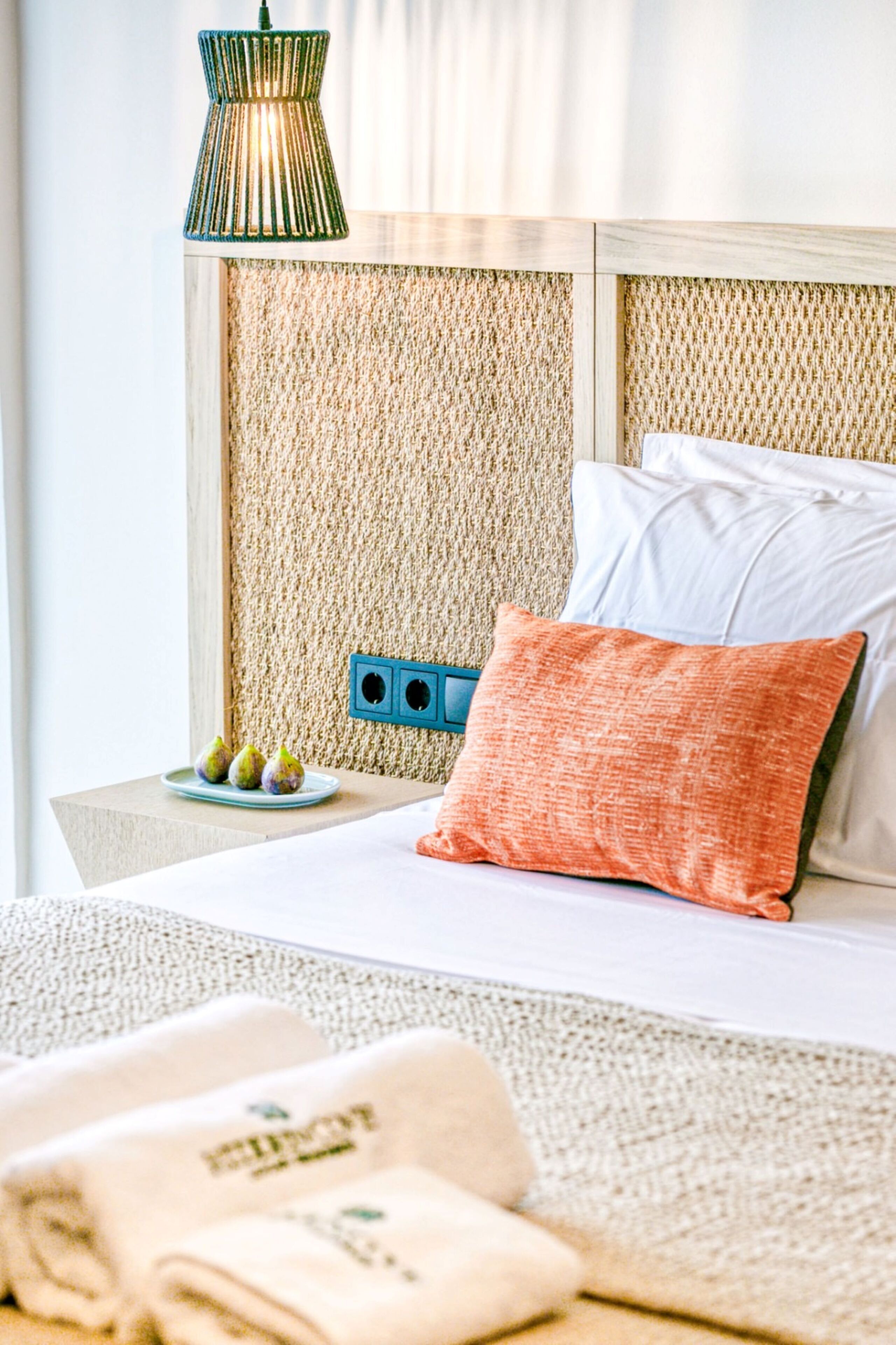Una cama bien hecha con un cojín blanco y naranja, una mesita de noche azul con una bandeja de fruta, y una pared con textura detrás de la cama.