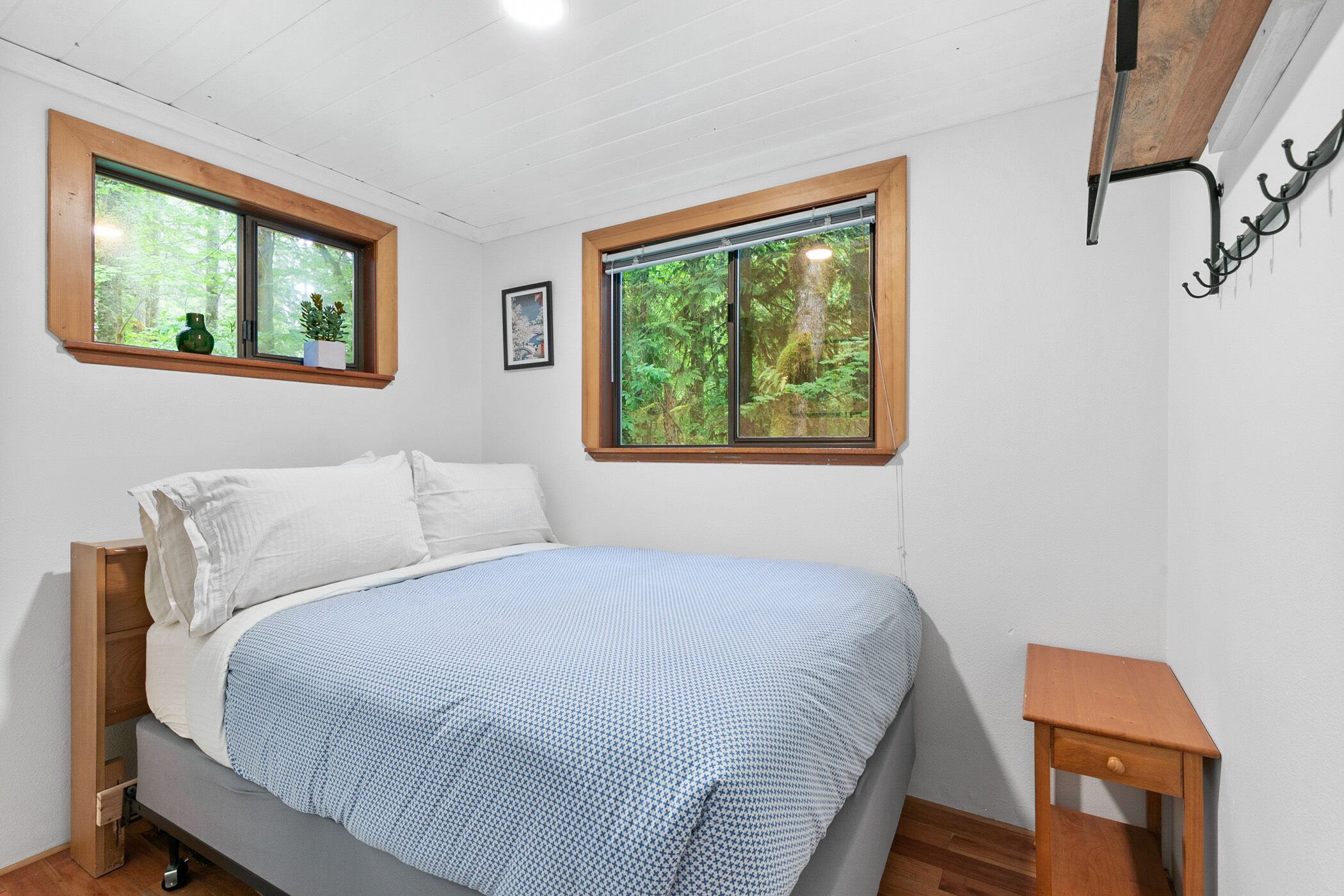 A bedroom with a bed, two windows with wooden frames, a small bedside table, and a wall-mounted hook.