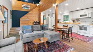 Un intérieur moderne avec un canapé d’angle gris, des poutres en bois, une cuisine aux meubles blancs et un tapis à motifs rouges.