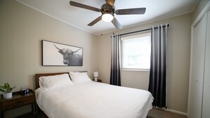 Un letto rifatto con lenzuola bianche, una ventola a soffitto, un comodino con una lampada, una sveglia e un quadro appeso alla parete.