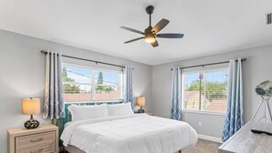 A bedroom with a bed, two bedside tables, a ceiling fan, and large windows with curtains.