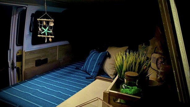Een compact, goed verlicht slaapgedeelte met een bed, kussens en een klein tafeltje met een plant.