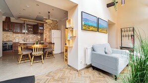 A modern kitchen with wooden cabinets, a dining area with a wooden table and chairs, a gray sofa, and a large window with a view of a landscape.