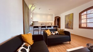 A modern living room with two black sofas, a wooden floor, and a kitchen in the background. - Apartment Alegría | Stylish accommodation right by the sea (Playa Honda)