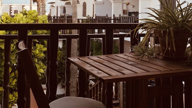 A wooden table and chair on a balcony with a view of a white building and palm trees. - Apartment Alegría | Stylish accommodation right by the sea (Playa Honda)