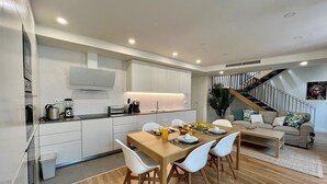 Una cocina moderna con armarios blancos, un comedor con una mesa de madera y sillas blancas, y una escalera que lleva a un dormitorio tipo loft.