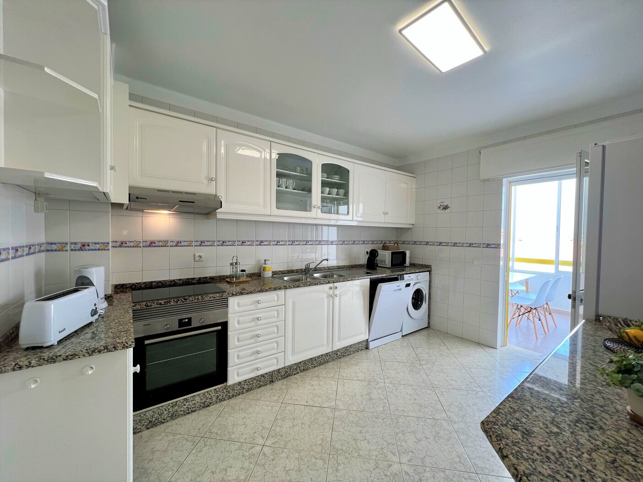 Une cuisine moderne avec des meubles blancs, des plans de travail en granit, un four encastré, un four à micro-ondes et un lave-linge.