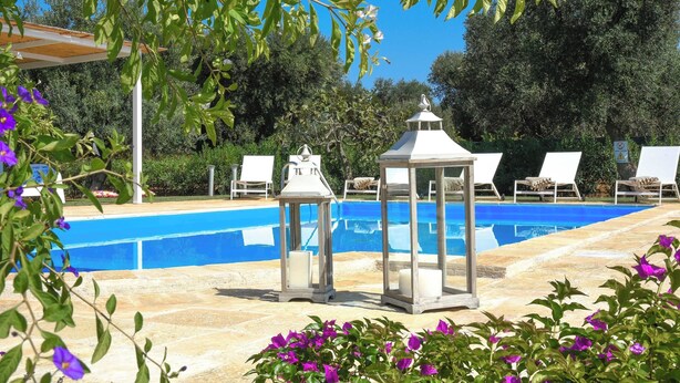 Una piscina con sillas de descanso y faroles, rodeada de follaje y flores.