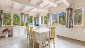 Un comedor con una mesa y sillas blancas, ventanas grandes con cortinas y vistas a árboles.