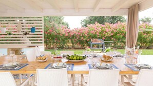 Un área de comedor al aire libre con una mesa de madera lista para una comida, rodeada de sillas blancas, con un jardín y flores rosadas al fondo.